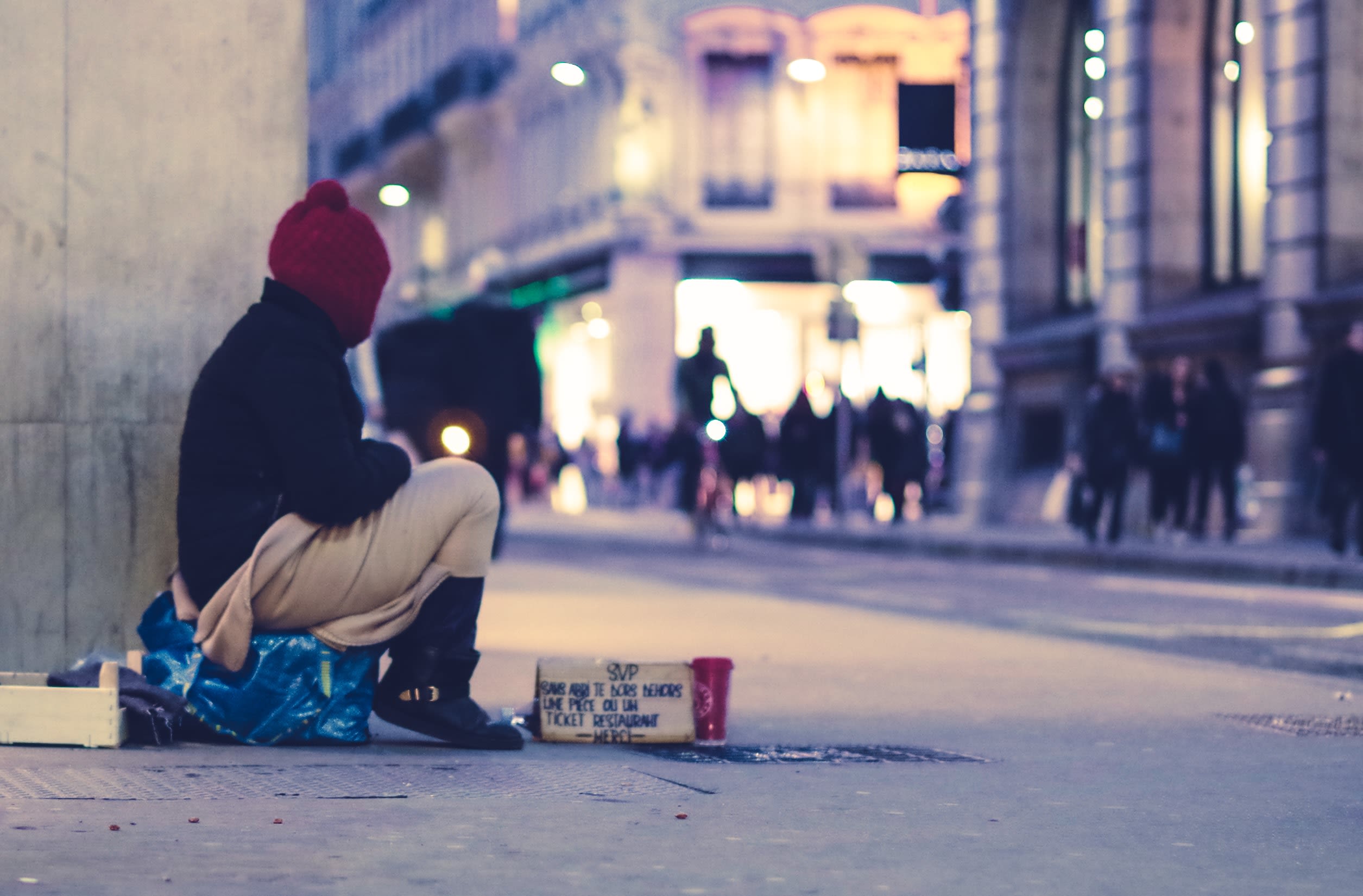 An unhoused person sitting at the corner of a building looking at a populated street.