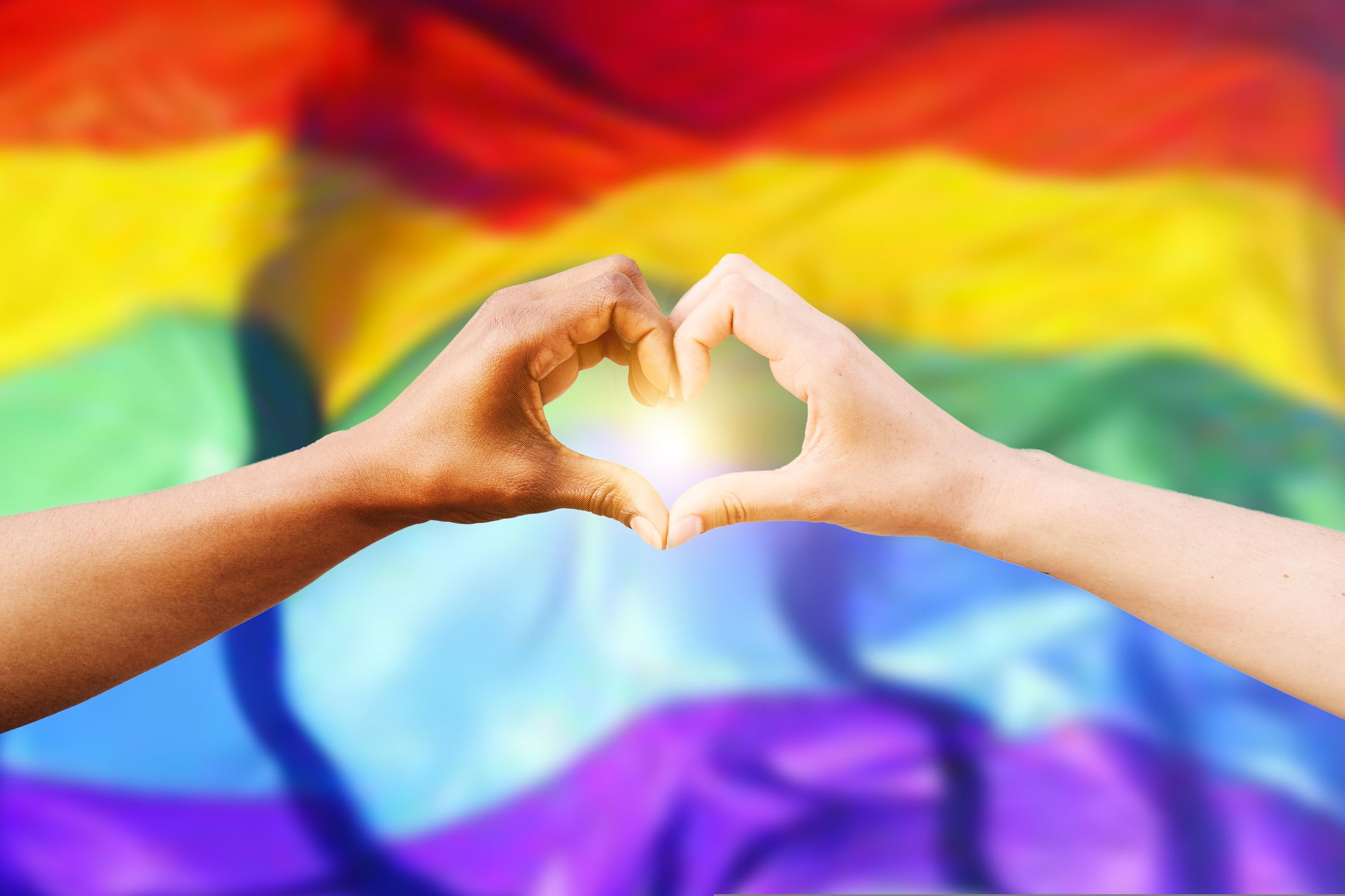 Two hands forming a heart in front of a Pride flag.